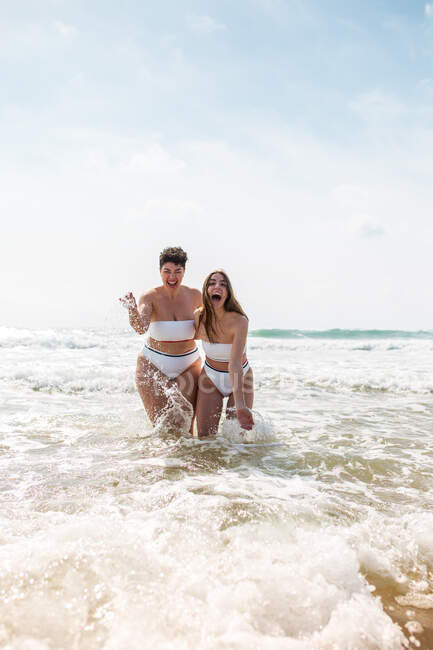 Веселі друзі-жінки в купальниках обіймаються один з одним, стоячи бризкаючи воду в піщаному океані біля піщаного пляжу під блакитним хмарним небом в сонячний день — стокове фото
