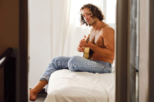 Seitenansicht eines talentierten männlichen Musikers mit nacktem Oberkörper und lockigem Haar, der auf dem Bett sitzt und Ukulele spielt — Stockfoto