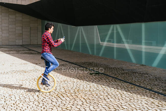Вид збоку молодого чоловіка в повсякденному вбранні переглядаючи смартфон під час їзди на одноколісному велосипеді на тротуарі біля сучасної будівлі з геометричним фасадом — стокове фото