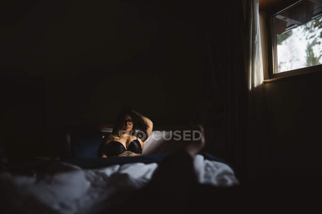 Joven mujer pensativa tatuada en lencería descansando en la cama en la oscuridad - foto de stock