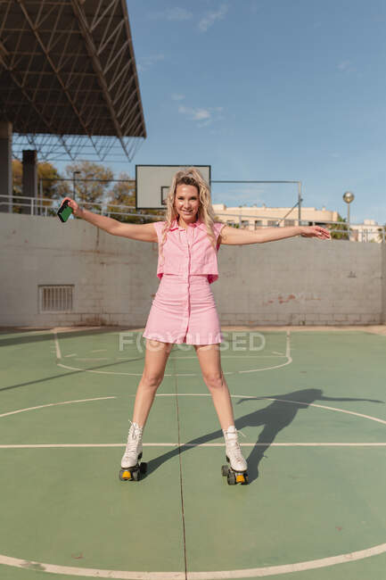 Comprimento total feminino ajuste alegre em rosa sundress patinação em rolos com braço levantado e tirar fotos na câmera de fotos instantâneas no chão de esportes ensolarado — Fotografia de Stock