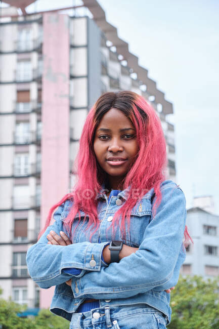 Selbstbewusste afroamerikanische Jugendliche mit rosa gefärbten Haaren tragen Jeanskleidung und schauen in die Kamera, während sie vor städtischen Gebäuden stehen — Stockfoto