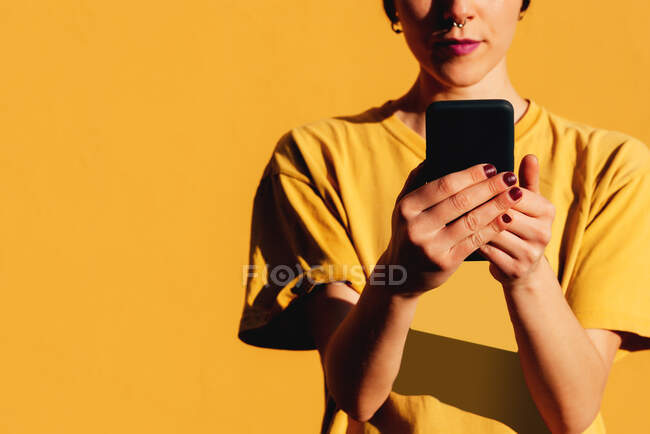 Современная женщина со стильной стрижкой и пирсингом с помощью смартфона в социальных сетях на жёлтом фоне — стоковое фото