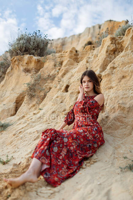 Спокойная женщина в длинном летнем платье сидит на песчаном холме и смотрит в камеру — стоковое фото