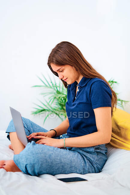 Positive junge Freiberuflerin in lässiger Kleidung sitzt mit gekreuzten Beinen auf dem Bett und tippt auf dem Laptop, während sie zu Hause an einem Remote-Projekt arbeitet — Stockfoto