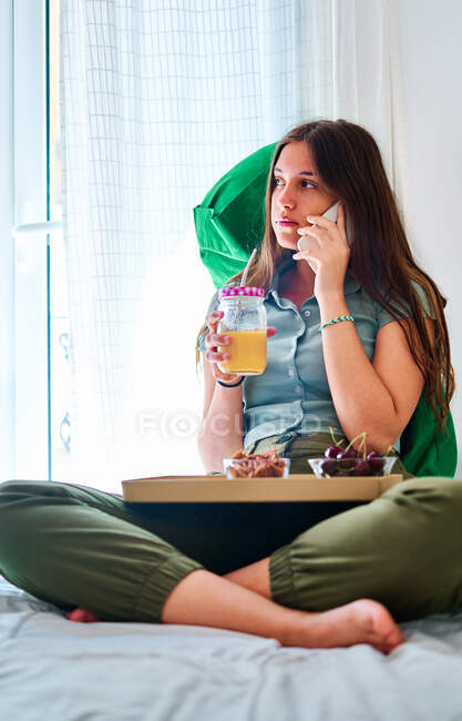 Young female student speaking on mobile phone near table with fresh fruits and juice while spending morning at home — Stock Photo