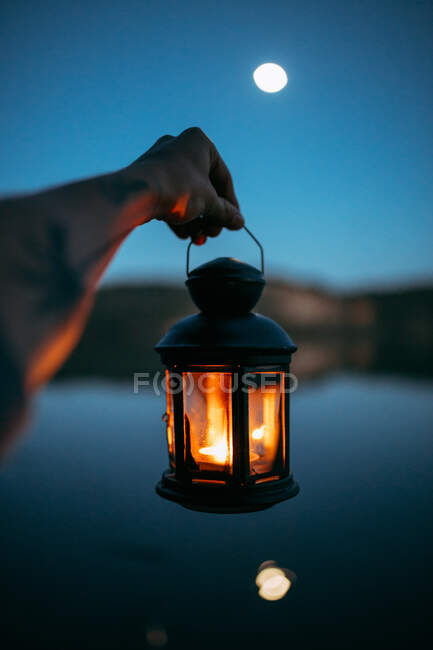 Crop hand of person holding chandelier with burning candle near water surface and moon in sky at night on blurred background — Stock Photo