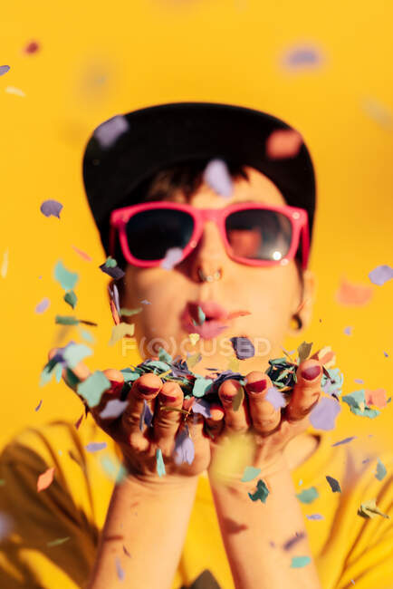 Сучасна жінка в хіп-хоп шапці і сонцезахисних окулярах, що дме барвистий конфетті і розважається на жовтому тлі — стокове фото