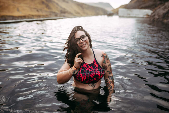 Joven hipster tatuado alegre en traje de baño en el agua entre las montañas - foto de stock