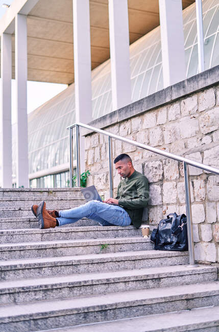 Hispanic man with coffee to go browsing data on netbook while sitting on steps and working on remote project on city street — Stock Photo