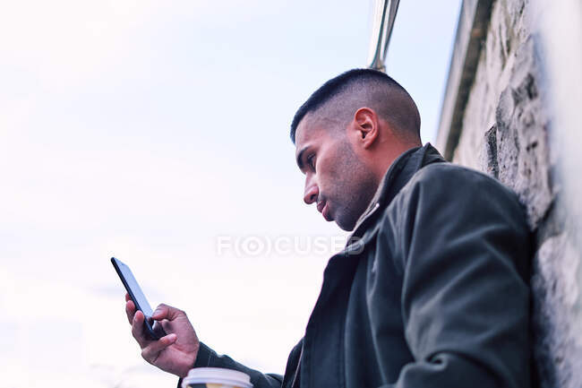Low angle of glad Hispanic man with coffee to go while leaning on stone wall and speaking on cellphone on street — Stock Photo