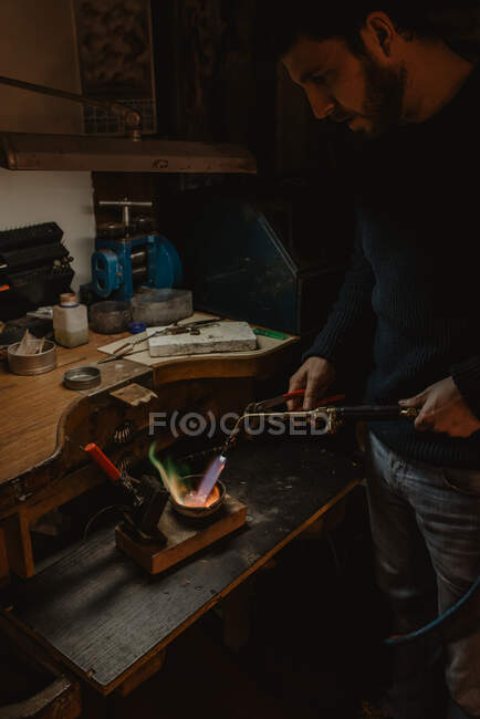 Goldsmith melting metal for jewellery with blowtorch while standing near workbench in workshop — Stock Photo