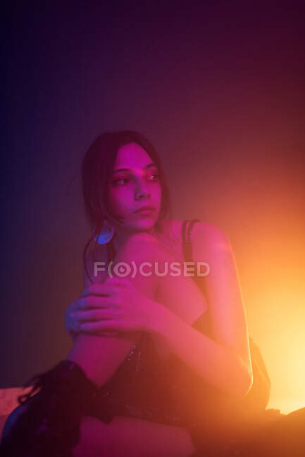 Tranquil young female model in dress sitting on floor and leaning on knee while looking away in dark studio with colorful lights — Stock Photo
