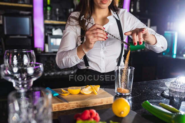 Cropped unrecognizable female barkeeper in stylish outfit adding sugar into glass while preparing mojito cocktail standing at counter in modern bar — Stock Photo