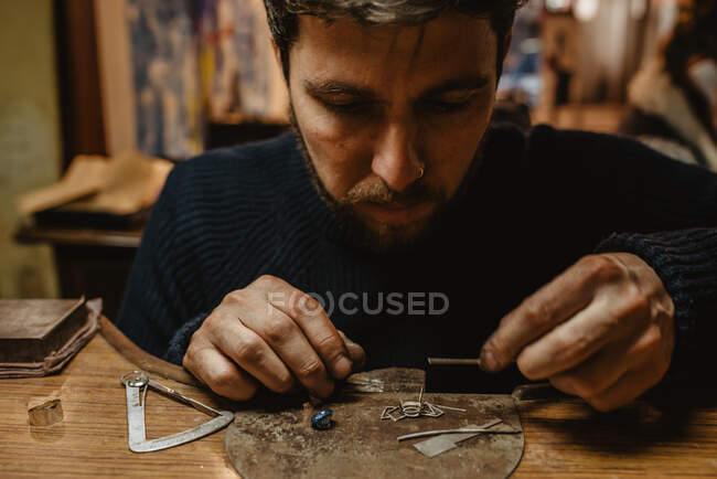 Бородатый мужчина ювелир, используя щипцы, делая крошечные металлические детали на верстаке в мастерской — стоковое фото