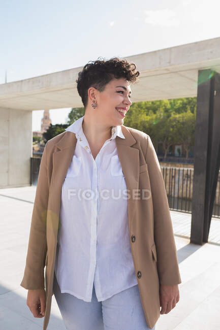 Joyeux jeune femme avec les cheveux courts dans des vêtements à la mode debout sur la passerelle dans la ville regardant loin dans la journée ensoleillée d'été — Photo de stock