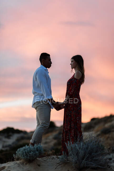 Amante pareja multirracial en ropa elegante abrazando en la colina en el fondo del cielo al atardecer sobre el mar en verano - foto de stock