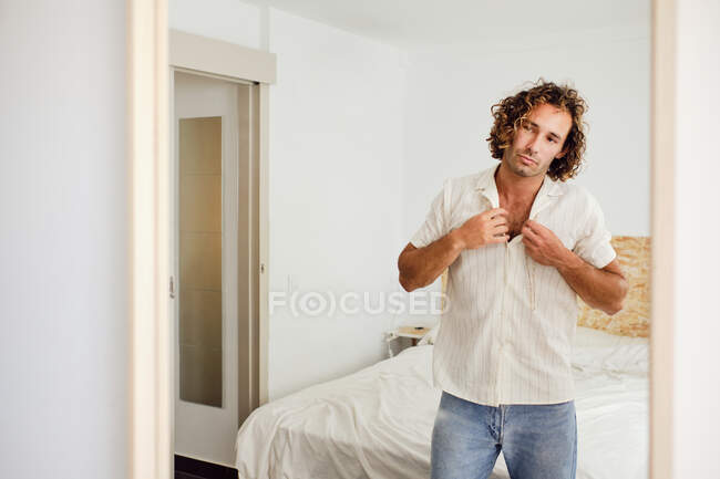 Riflessione di elegante maschio con capelli ricci abbottonatura camicia mentre si guarda nello specchio in camera — Foto stock