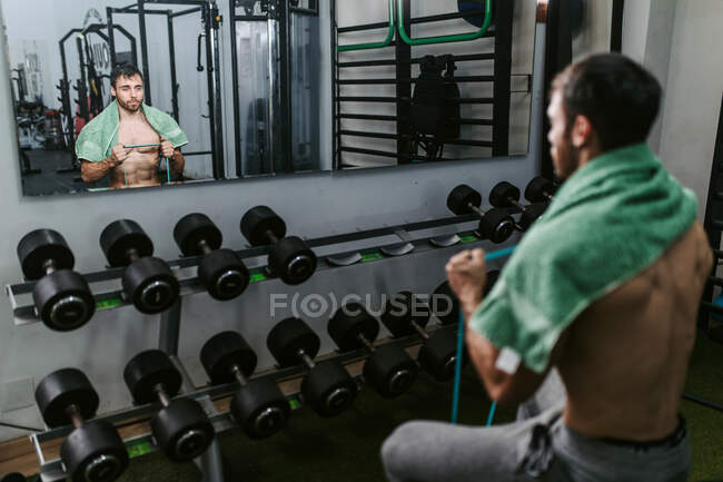 Shirtless man sitting on a bench stretching elastic band with arms during functional workout in gym — Stock Photo