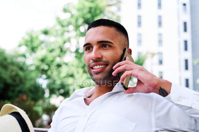 Bajo ángulo de sonriente auto seguro joven barbudo caballero hispano en traje elegante y sombrero hablando en el teléfono móvil mientras descansa en el banco en la ciudad - foto de stock