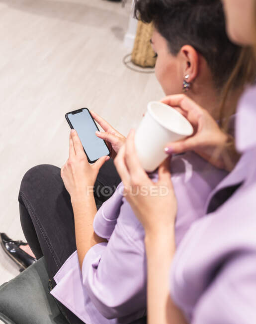 Обрезание анонимных женщин в модном наряде просматривает по мобильному телефону в светлой комнате рядом со стулом — стоковое фото