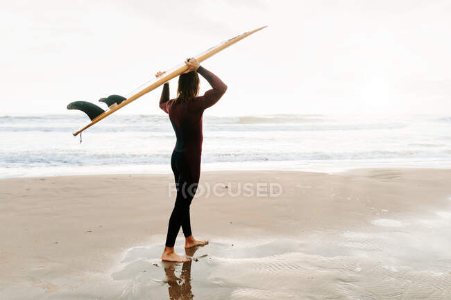 Vista laterale di un giovane surfista irriconoscibile con i capelli lunghi vestito con muta in piedi guardando lontano sulla spiaggia con la tavola da surf sulla testa durante l'alba — Foto stock