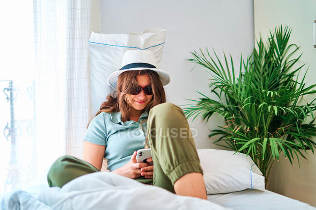 Smiling young female in summer wear and hat with sunglasses sitting on bed and browsing on smartphone while spending time alone at home — Stock Photo