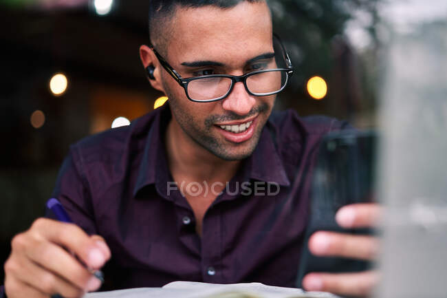 Bello giovane imprenditore ispanico di sesso maschile che controlla le informazioni su smartphone e scrive note in agenda mentre lavora a tavola con il computer portatile — Foto stock