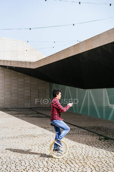 Side view of young male in casual outfit browsing on smartphone while riding unicycle on pavement near modern building with geometric facade — Stock Photo