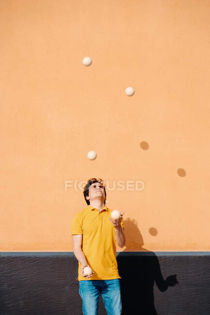 Joven talentoso macho realizando truco con bolas malabares mientras está de pie en el pavimento cerca de la pared naranja brillante - foto de stock
