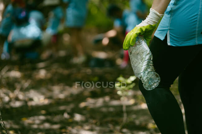 Cultive ativista anônimo em luvas de proteção pegando garrafa de plástico do chão durante a coleta de lixo na natureza — Fotografia de Stock