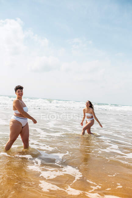 Vista lateral de amigas en trajes de baño en el océano espumoso cerca de la playa de arena bajo el cielo azul nublado en el día soleado - foto de stock