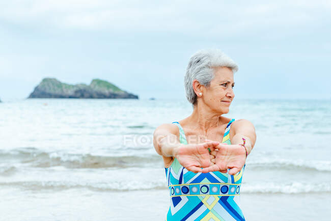 Active old gray haired female pensioner in stylish swimsuit stretching arms and shoulders while doing exercises on beach against waving ocean — Stock Photo