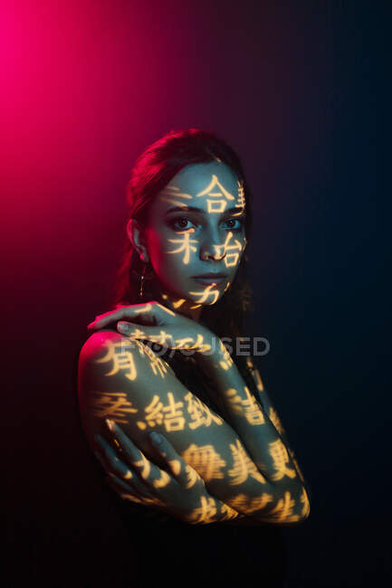 Modello femminile giovane alla moda con proiezione di luce a forma di geroglifici orientali che guardano la macchina fotografica in studio scuro con illuminazione rossa — Foto stock