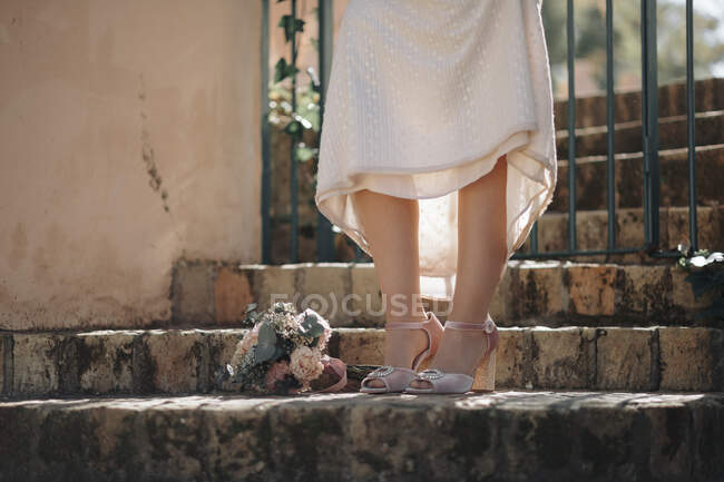 Crop anonymous bride in white wedding dress and pink high heeled peep toe shoes standing near bridal bouquet on weathered stone stairs — Stock Photo