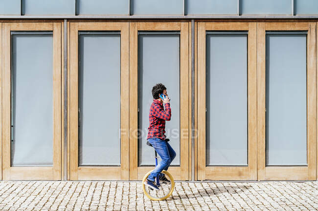 Full body side view of young male in casual outfit answering phone call while riding unicycle on pavement near modern building with geometric facade — Stock Photo