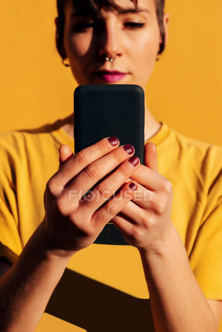 Mujer contemporánea con corte de pelo elegante y piercing usando smartphone en las redes sociales contra el fondo amarillo - foto de stock