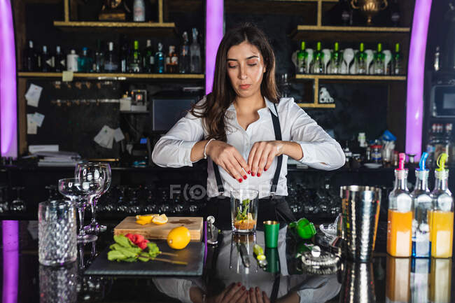 Junge Barkeeperin in stylischem Outfit bereitet Mojito-Cocktail mit Zitronenscheiben zu, während sie an der Theke in einer modernen Bar steht — Stockfoto