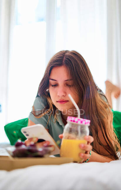 Junge Studentin surft in sozialen Netzwerken auf dem Handy am Tisch mit frischem Obst und Saft, während sie den Morgen zu Hause verbringt — Stockfoto