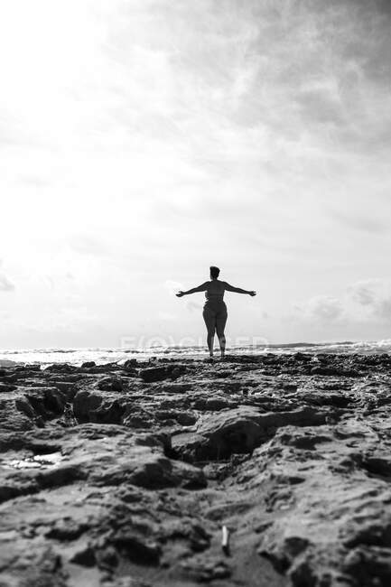 Rückansicht einer gesichtslosen Frau in Badebekleidung, die mit erhobenen Händen an der felsigen Küste mit Felsbrocken in der Nähe des Meeres am Tag unter dem Himmel steht — Stockfoto
