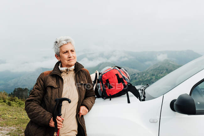 Elderly woman with walking stick leaning on white vehicle and looking away against mountain during road trip in nature — Stock Photo