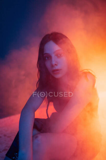 Tranquillo giovane modello femminile in abito seduto sul pavimento guardando la fotocamera in studio scuro con luci colorate — Foto stock