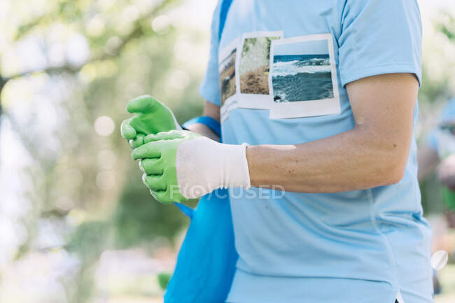 Activista anónimo de cultivos en camisa azul y guantes de goma verde que lleva bolsa de basura mientras recoge basura en el parque verde en el día de verano - foto de stock