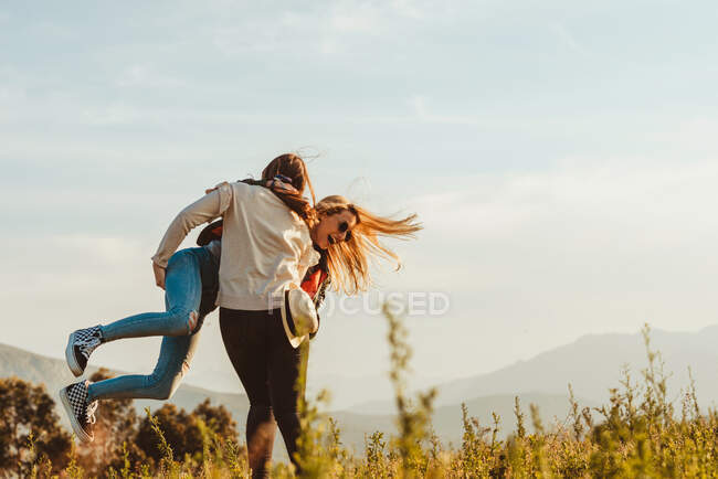 Обратный вид женщины, несущей взволнованную подружку на руках, кружащуюся вокруг, стоя на поле в горах — стоковое фото