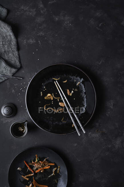 Overhead black empty bowl with pair of chopsticks and leftovers of Korean cuisine Japchae at table near full plate — Stock Photo