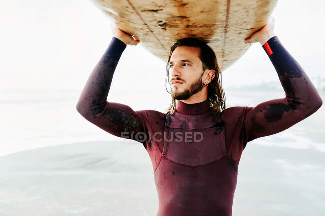 Retrato de un joven surfista con el pelo largo y la barba vestida con traje de neopreno de pie mirando hacia la playa con la tabla de surf sobre la cabeza durante el amanecer - foto de stock