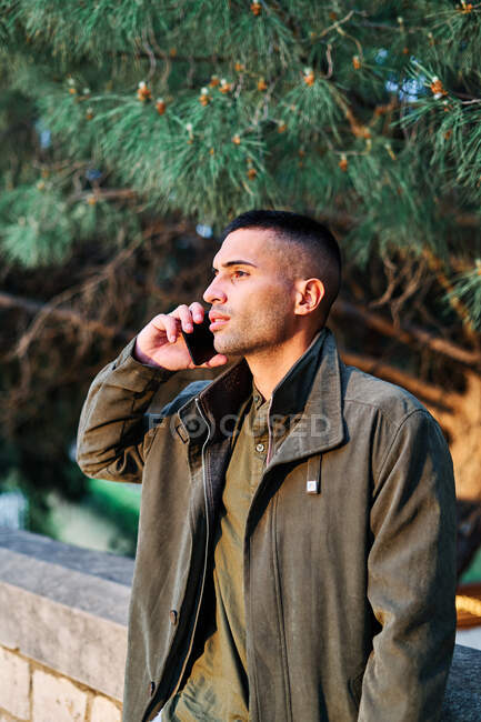 Hispânico cara em roupas casuais olhando para longe e atender telefonema enquanto estava perto de barreira e árvore conífera no parque — Fotografia de Stock
