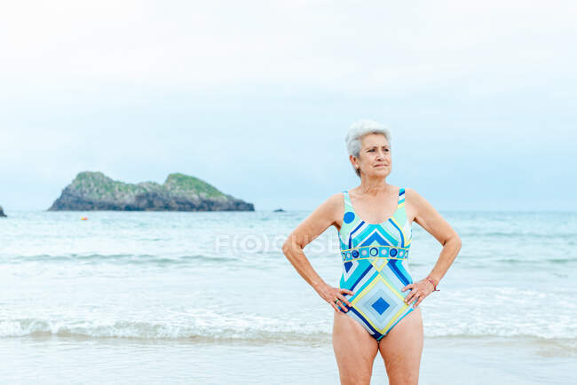 Ältere Frau mit durchtrainiertem Körper trägt einen eleganten, farbenfrohen Badeanzug mit geometrischem Print und steht mit den Händen an der Taille an einem Sommertag gegen das Meer — Stockfoto