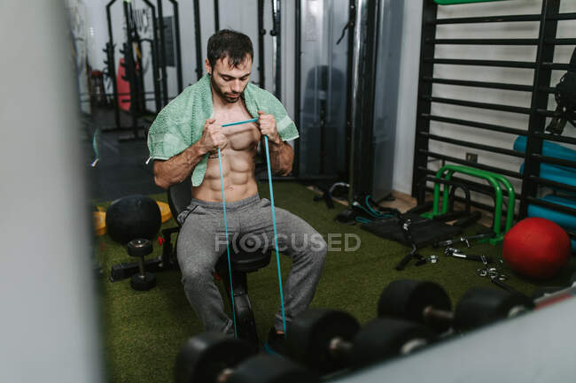 Мужчина без рубашки сидит на скамейке, растягивая эластичную ленту с руками во время функциональной тренировки в спортзале — стоковое фото