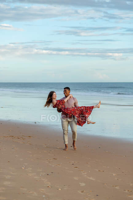 Feliz hombre negro llevando a la mujer encantada y disfrutando del verano en la orilla del mar de arena en el fondo del mar al atardecer - foto de stock